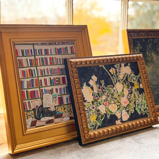 Two framed artworks, one floral still life and one colorful library scene, on a marble surface with a window in the background